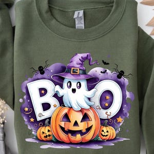 Puede incluir: Sudadera verde oliva con un diseño de Halloween. El diseño presenta un fantasma de dibujos animados con un sombrero de bruja, sentado en una calabaza, con la palabra "BOO". Arañas, murciélagos y estrellas completan el diseño.
