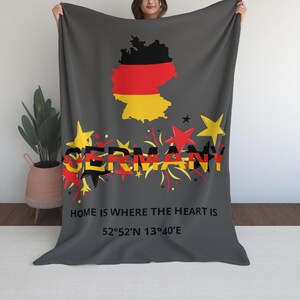 Puede incluir: Una manta gris oscuro con un mapa de Alemania en los colores de la bandera alemana. La palabra "GERMANY" se muestra en una fuente estilizada, junto con la frase "HOME IS WHERE THE HEART IS" y coordenadas.