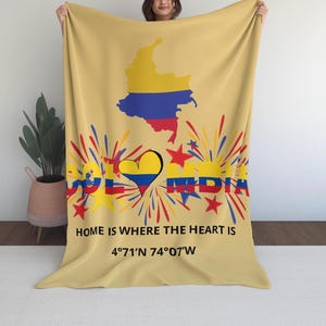 Puede incluir: Una manta de color amarillo claro con un mapa de Colombia en amarillo, azul y rojo. La manta muestra la palabra "COLOMBIA" en rojo, amarillo y azul, junto con un corazón y estrellas. También se ve el texto "HOME IS WHERE THE HEART IS" y las coordenadas.