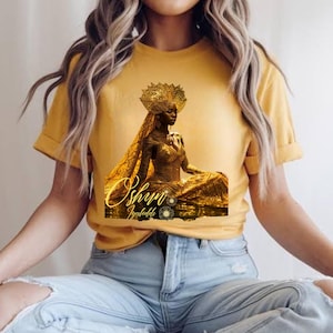 Op de afbeelding: Mosterdgeel T-shirt met een gouden illustratie van een gekroonde figuur in een vloeiende jurk. De tekst "Oshun Iyaledde" is inbegrepen. Het T-shirt is gemaakt van een zacht materiaal. Een casual kledingstuk.