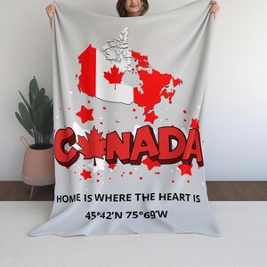 Puede incluir: Una manta gris claro con un mapa rojo y blanco de Canadá, la palabra "CANADA" en letras rojas en 3D y acentos de estrellas rojas. También se ve el texto "HOME IS WHERE THE HEART IS" y las coordenadas.
