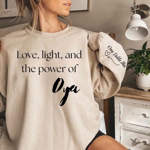 Può includere: Una felpa beige con la scritta "Love, light, and the power of Oya" in nero. La manica ha la scritta "Omo Ladde Iku" in nero. La felpa è di colore neutro.