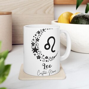 Puede incluir: Taza de cerámica blanca con un símbolo del zodiaco Leo negro, un diseño de media luna con estrellas y flores, y el texto "Leo Custom Name". La taza está sobre un posavasos beige.