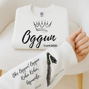 Puede incluir: Sudadera blanca con texto negro y diseño de corona. El texto incluye "Oggun" y "Est 2026". La manga presenta texto y un gráfico de una espada. La sudadera está doblada sobre una silla blanca.