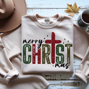 Puede incluir: Sudadera color crema con las palabras "merry CHRISTmas" en una fuente desgastada. La cruz es roja y las letras son verdes y rojas. Un sombrero marrón y una taza de café están en el fondo.