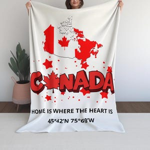 Puede incluir: Manta blanca con un mapa rojo de Canadá, diseños de hojas de arce y la palabra "CANADA" en rojo. El texto "HOME IS WHERE THE HEART IS" y las coordenadas también se muestran.