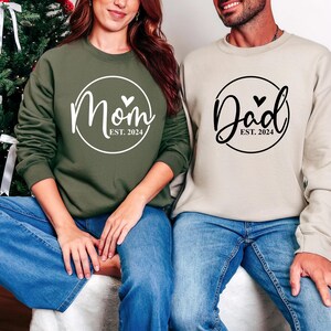 Puede incluir: Dos sudaderas de manga larga, una verde oliva y otra beige, con diseños a juego. La sudadera verde oliva dice "Mom Est. 2024" en escritura blanca, y la sudadera beige dice "Dad Est. 2024" en escritura negra. Ambos diseños incluyen un corazón.