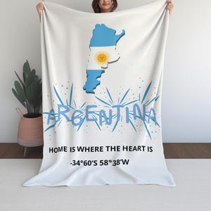 Puede incluir: Manta blanca con un mapa azul y blanco de Argentina y un emblema de sol. La palabra "ARGENTINA" está escrita en azul con una fuente de estilo grafiti. También se muestra el texto "HOME IS WHERE THE HEART IS" y las coordenadas.