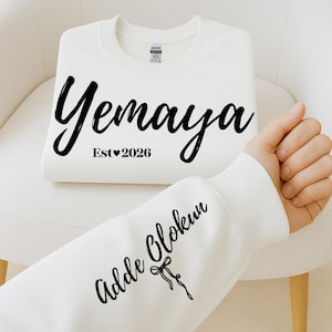 Puede incluir: Una sudadera blanca con el nombre "Yemaya" en fuente negra. Debajo, "Est 2026" con un corazón. La manga muestra "Adde Olokun" en fuente negra con un lazo y una cinta.