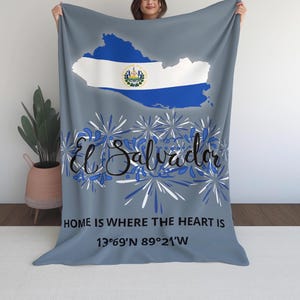 Puede incluir: Una manta gris con el contorno de El Salvador y su bandera. La manta tiene el texto "El Salvador", "HOME IS WHERE THE HEART IS" y las coordenadas 13°69'N 89°21'W. Acentos florales azules y blancos rodean el texto.