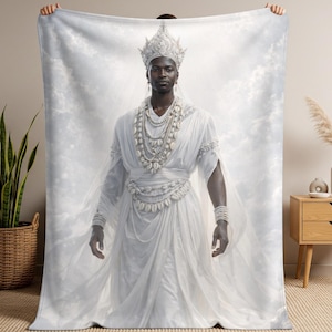 May include: A rectangular throw blanket featuring a printed image of a person wearing a white robe, a tall crown, and multiple layered shell necklaces. The background of the print consists of white clouds and soft light, creating a bright aesthetic.