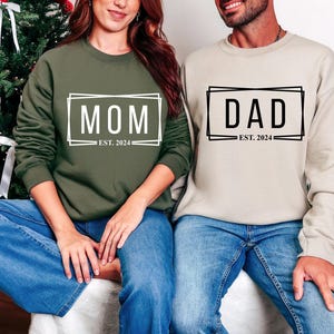 Puede incluir: Dos personas con sudaderas a juego. Una es verde oliva con la palabra "MOM" y "EST. 2024" en un diseño rectangular blanco. La otra es beige con "DAD" y "EST. 2024" en un diseño rectangular negro.