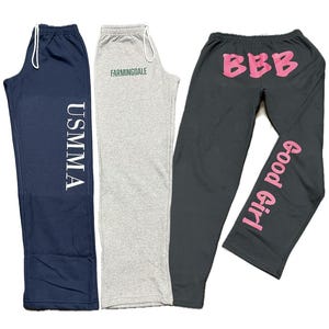 May include: Three pairs of sweatpants in navy blue, gray, and charcoal. The navy pants have "USMMA" printed vertically in white. The gray pants have "Farmingdale" printed in green. The charcoal pants have "BBB" and "Good Girl" printed in pink.