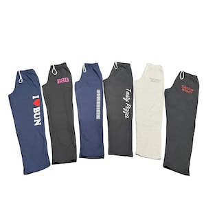 Może przedstawiać: Sześć par spodni dresowych w różnych kolorach, każda z talią ze sznurkiem i unikalnym tekstem. Spodnie są granatowe, czarne i szare, z nadrukami takimi jak "I BUN", "BBQ" i "Tasty Pizza".