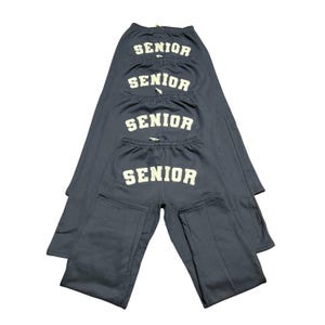 May include: Four pairs of navy blue sweatpants with the word "SENIOR" printed in white block letters across the back. The sweatpants have an elastic waistband and a relaxed fit. The image is against a white background.
