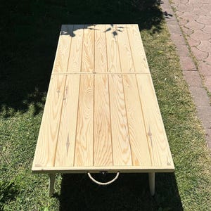 Peut inclure: Une table pliante rectangulaire en bois clair avec une poignée en corde. La table est faite de planches de bois verticales et possède quatre pieds courts. La table est posée sur une zone herbeuse.