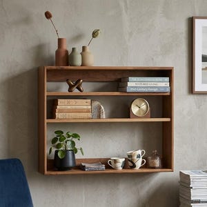 May include: A rectangular wooden wall shelf with three horizontal tiers. The shelves hold books titled The Scandinavian Home, Textured Living, and Mid-Century Design, a brass clock, a green plant in a black pot, ceramic mugs, and vases.