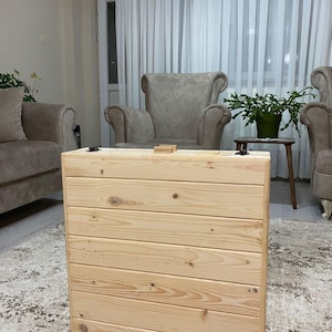 May include: A light-colored wooden storage chest with a hinged lid. The chest is made of horizontal wooden planks and has a natural wood grain finish. It is placed on a patterned rug in a living room setting.