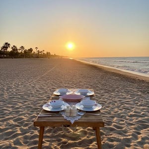May include: A wooden table set for a meal on a sandy beach at sunset. The table holds plates, bowls, silverware, and salt and pepper shakers. The sun is setting over the ocean, with palm trees in the distance.