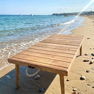 May include: A rectangular wooden table with a slatted top and four legs. The table has a natural wood finish and is positioned on a sandy beach. A rope handle is attached to the side. The background features the ocean and a clear blue sky.