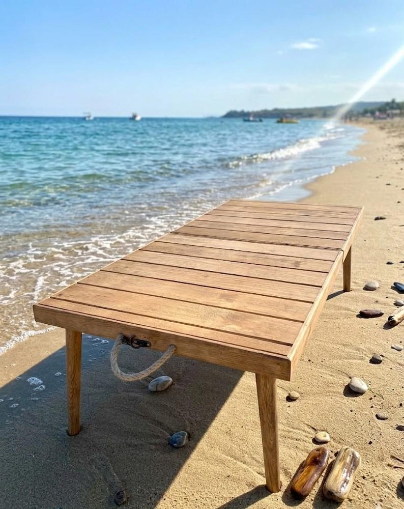 May include: A rectangular wooden table with a slatted top and four legs. The table has a natural wood finish and is positioned on a sandy beach. A rope handle is attached to the side. The background features the ocean and a clear blue sky.