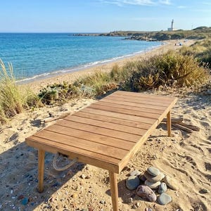 May include: A rectangular wooden table with a slatted top and four legs, set on a sandy beach. The table has a natural wood finish and a rope handle. The background shows a beach, ocean, and a lighthouse.