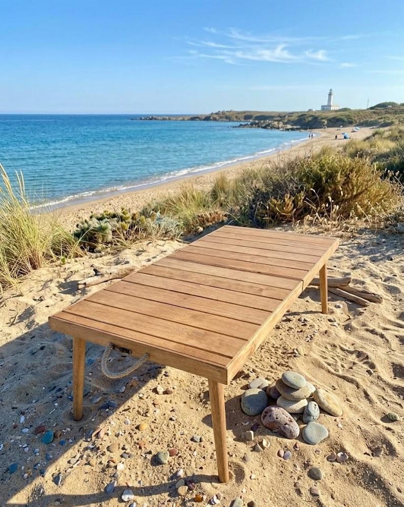 May include: A rectangular wooden table with a slatted top and four legs, set on a sandy beach. The table has a natural wood finish and a rope handle. The background shows a beach, ocean, and a lighthouse.