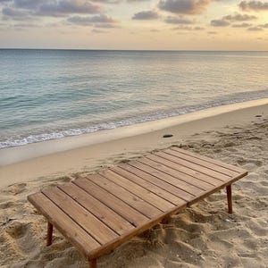 Może przedstawiać: Prostokątny drewniany stół z czterema nogami stoi na piaszczystej plaży. Stół wykonany jest z jasnobrązowego drewna i ma listwowy blat. W tle spokojne morze i zachmurzone niebo.