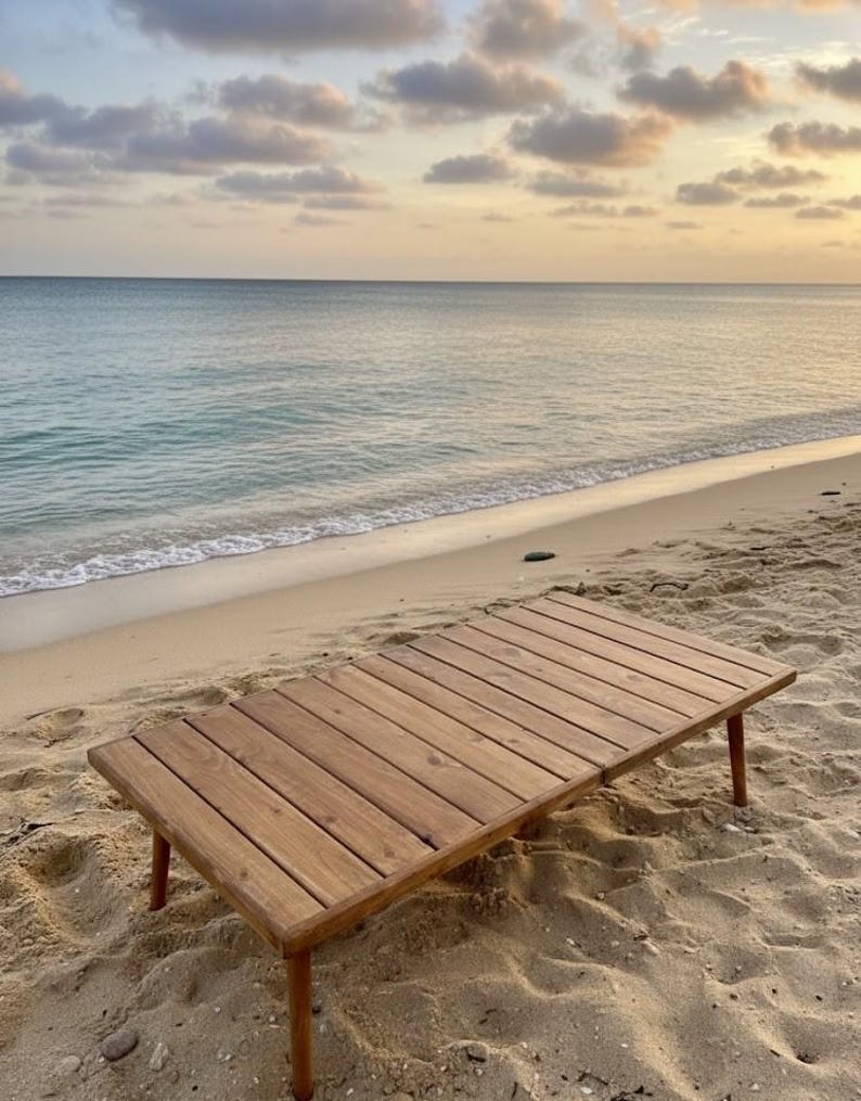 May include: A rectangular wooden table with four legs, constructed from light brown planks. The table is set on a sandy beach, with the ocean and a cloudy sky in the background. The table is approximately 48 inches long and 24 inches wide.