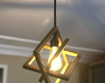 May include: A wooden pendant light fixture with a geometric design. The light fixture is made of multiple pieces of wood that are arranged in a square shape. The light fixture is hanging from a black cord.