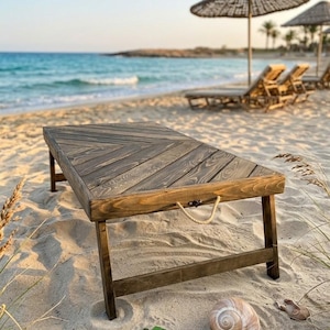 Op de afbeelding: Een houten strandtafel met een visgraatpatroon op de bovenkant, op een zandstrand. De tafel heeft een donkerbruine afwerking en wordt ondersteund door vier poten. Een touwgreep is aan de zijkant bevestigd. De strandscène omvat een zeeschelp en zeeglas.