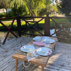 Op de afbeelding: Een houten tafel met een wit tafelkleed en een roze loper, gedekt voor vier personen met witte borden, kommen en bestek. De tafel staat op een houten dek met uitzicht op een klein beekje.
