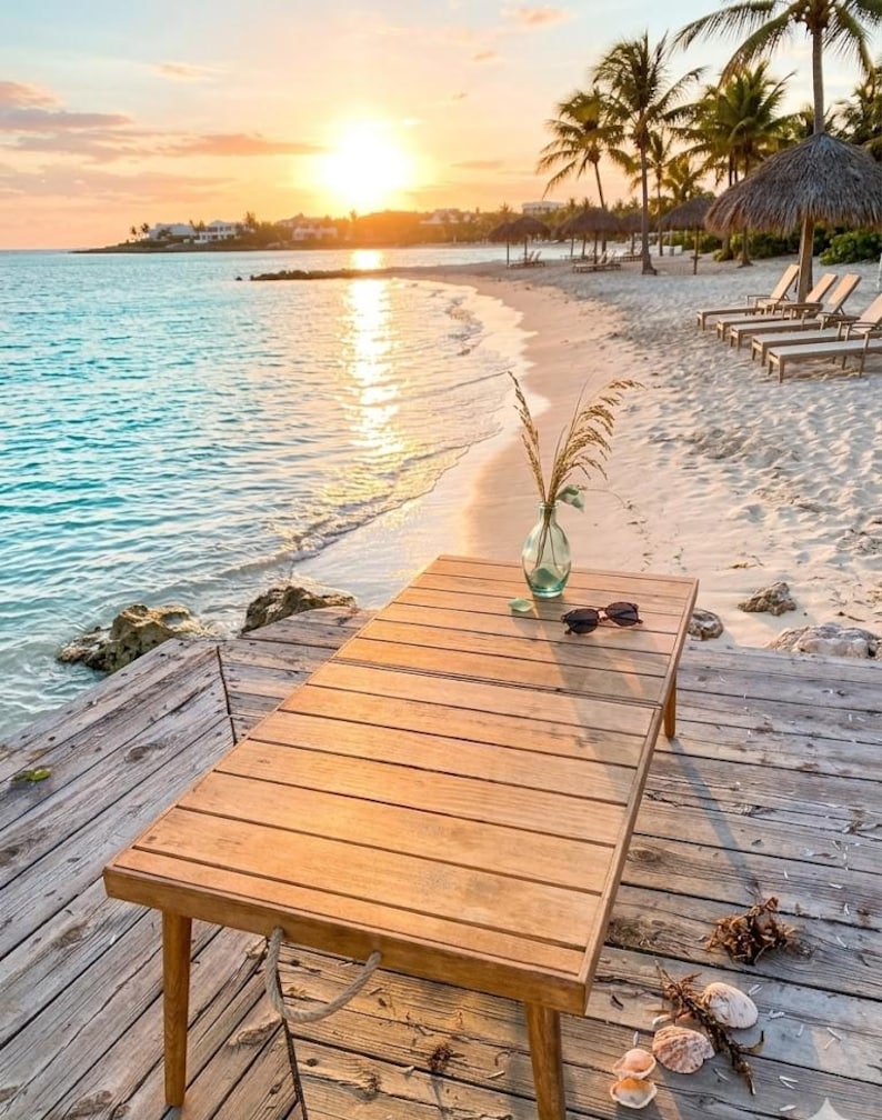May include: A wooden table with a vase of dried plants and sunglasses sits on a wooden dock. The beach scene includes a sandy shore, turquoise water, palm trees, and a sunset.