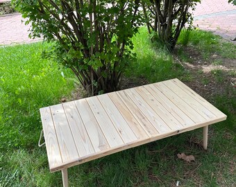 May include: A light wood folding table with a natural finish. The table is made of wooden planks and has two legs on each side. The table is sitting on a grassy surface.