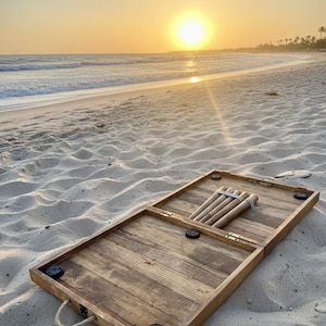 May include: A wooden backgammon set with a light brown finish, open on a sandy beach. The set includes wooden game pieces and black corner accents. The background features a sunset over the ocean.