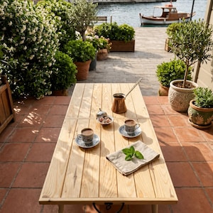 May include: A rectangular light-colored wooden coffee table with four legs and a black metal handle on the side. On the table are two patterned ceramic cups with dark coffee, a small silver bowl with Turkish delight, a copper pot, and a napkin.