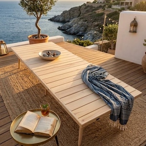May include: A low rectangular wooden table with horizontal slats, featuring a light natural finish. A blue and white patterned textile with fringe rests on the surface, alongside a shallow bowl containing smooth grey stones.
