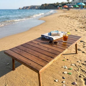 May include: A rectangular, dark brown wooden beach table with four legs, set on a sandy beach. The table holds a folded towel, a book, sunglasses, and a glass of iced tea with a lime wedge. The ocean and sky are visible in the background.