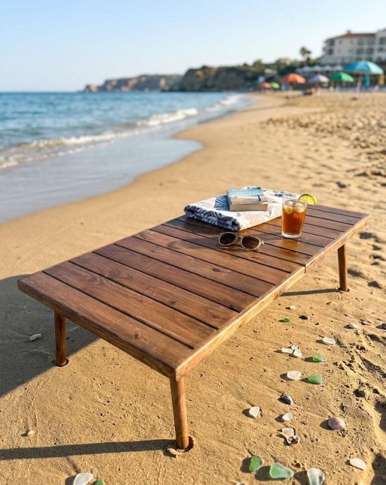 May include: A rectangular, dark brown wooden beach table with four legs, set on a sandy beach. The table holds a folded towel, a book, sunglasses, and a glass of iced tea with a lime wedge. The ocean and sky are visible in the background.