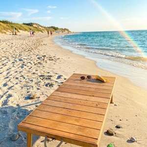 May include: A wooden beach table with sunglasses and sunscreen on a sandy beach. The table has a light brown color and is made of wooden planks. The beach has a blue ocean and a rainbow in the sky.