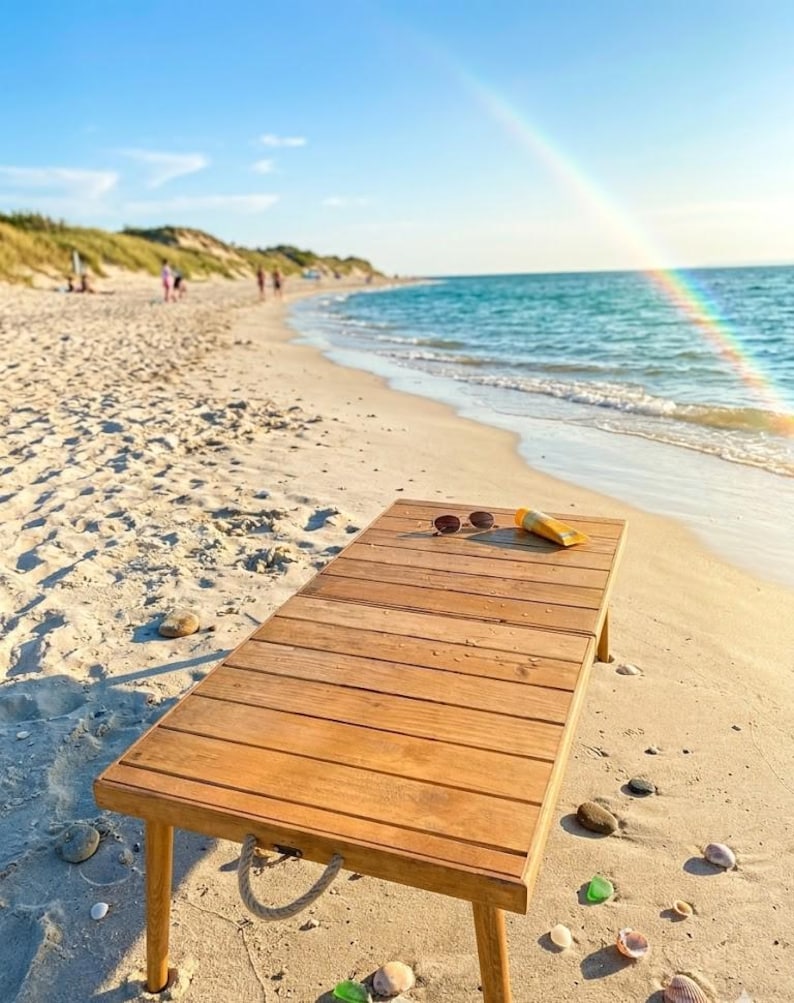 May include: A wooden beach table with sunglasses and sunscreen on a sandy beach. The table has a light brown color and is made of wooden planks. The beach has a blue ocean and a rainbow in the sky.