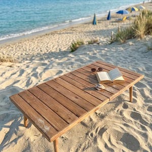 May include: A rectangular wooden table with a book and sunglasses on a sandy beach. The table is made of light brown wood and has four legs. The open book and sunglasses are on the table's surface.