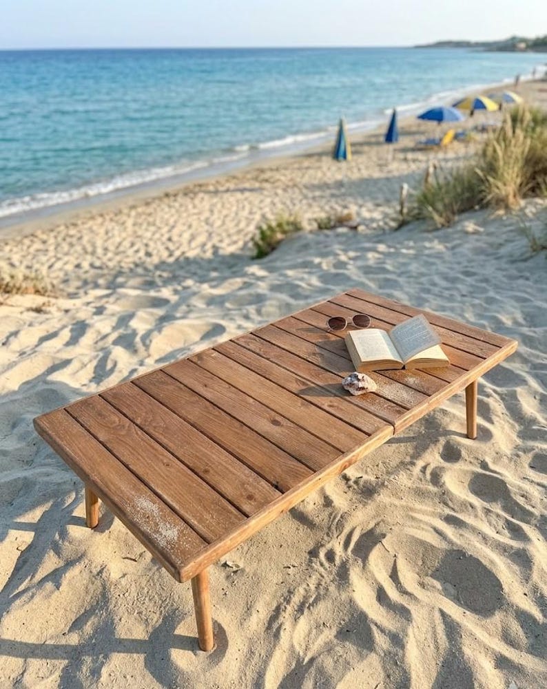 May include: A rectangular wooden table with a book and sunglasses on a sandy beach. The table is made of light brown wood and has four legs. The open book and sunglasses are on the table's surface.
