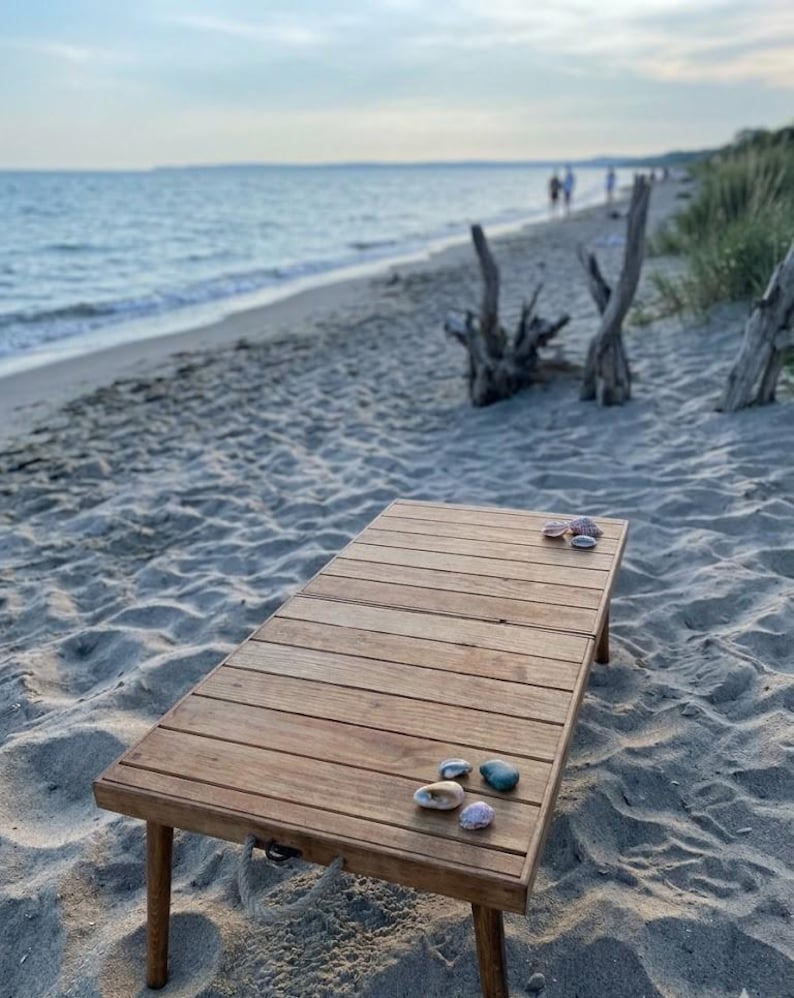 May include: A rectangular wooden table with four legs sits on a sandy beach. Several seashells and colorful stones are arranged on the table's surface. The background shows the ocean and a few people walking on the beach.