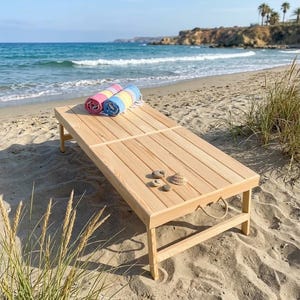 May include: A rectangular wooden beach lounger with a slatted surface, four legs, and a rope handle on the side. Two rolled-up striped towels in pink, yellow, and blue rest on the surface alongside three small smooth stones and a seashell.
