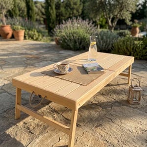May include: A rectangular light-colored wooden folding table with a slatted top and rope handles on the sides. A burlap placemat holds a ceramic teacup and saucer, a glass carafe with a lemon slice, and a book. A small wooden lantern sits nearby.