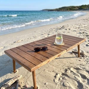 May include: A low, rectangular wooden table with folding legs sits on a sandy beach. Sunglasses rest on the table's surface, and a glass of water with lemon and mint sits on a coaster. The ocean and blue sky are in the background.