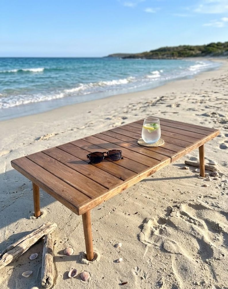 May include: A low, rectangular wooden table with folding legs sits on a sandy beach. Sunglasses rest on the table's surface, and a glass of water with lemon and mint sits on a coaster. The ocean and blue sky are in the background.