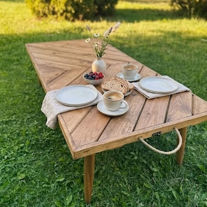 Puede incluir: Una mesa de picnic de madera preparada para una comida al aire libre. La mesa es de madera marrón claro y tiene un patrón de espiga. Está puesta con platos, tazas de café, pan y un pequeño jarrón con flores. La mesa tiene un asa de cuerda.