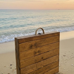 May include: A wooden folding table with a rope handle stands on a sandy beach. The table is constructed of horizontal wooden planks, stained a warm brown. The background features the ocean and a sunset.